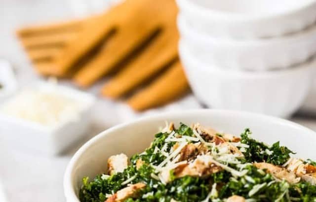 bowl of grilled chicken kale salad sprinkled with parmesan cheese with wood salad tongs