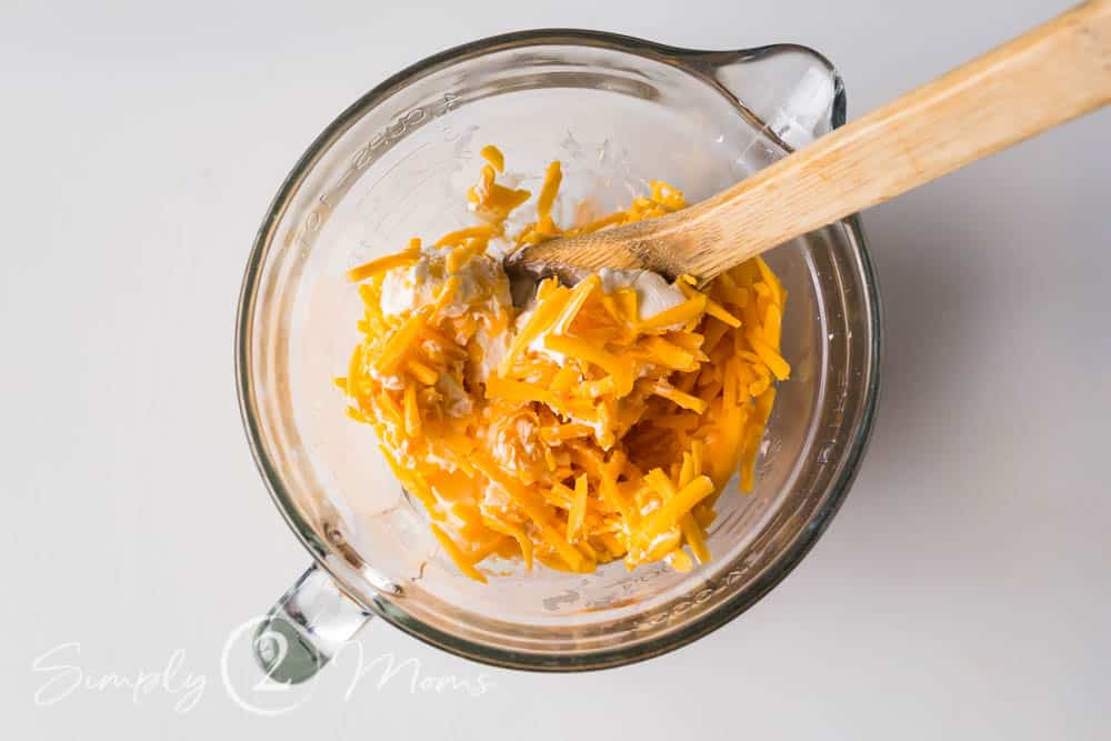 grated cheddar cheese and cream cheese in a clear glass mixing bowl with a wooden spoon