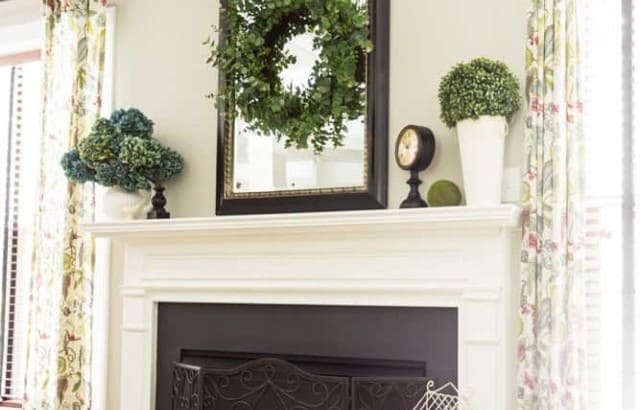 Summer fireplace decor ideas on a traditional gas fireplace with painted white mantel. Mantel has a pitcher of blue hydrangeas, large boxwood orb, large black mirror with succulent wreath layered on top. Small stool with white vintage birdcage on top next to fireplace on hearth. Rope basket with topiary orb on other side.