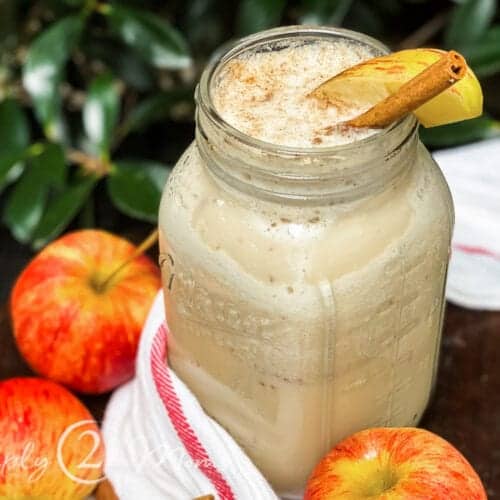 Overhead view of a large mason jar with a protein shake garnished with a slice of apple and a stick of cinnamon sitting on a striped piece of fabric surrounded by several apples and a stack of cinnamon sticks on a wood table with leaves in the background
