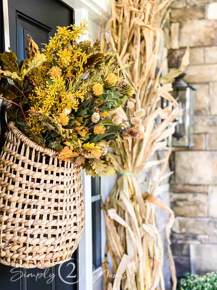 Target hyacinth door basket filled with gold faux fall florals hanging on a door with dried corn stalks leaning against stacked stone home exterior