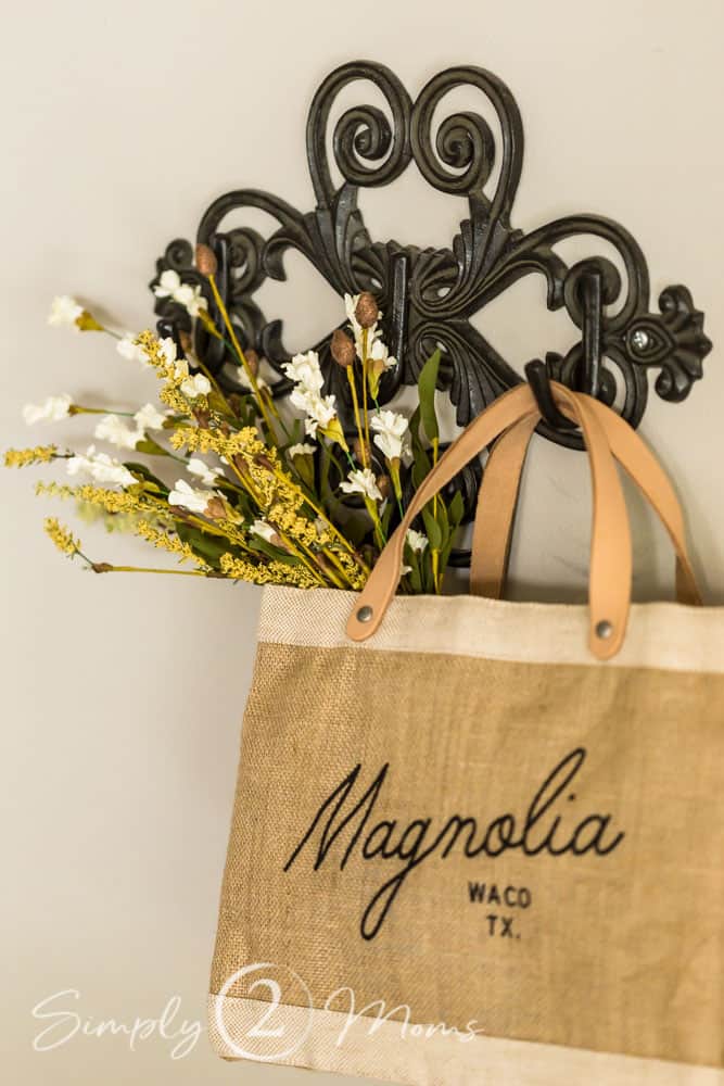 A Magnolia brand canvas shopping tote with leather handles is filled with dried white fall flowers and hangs from an iron scrollwork wall hanger.