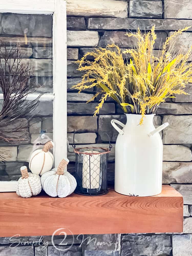 Stacked stone fireplace with antique window frame on wood mantel. Mantle has enamel jug with fall grasses to add fall color and three small sweater pumpkins.