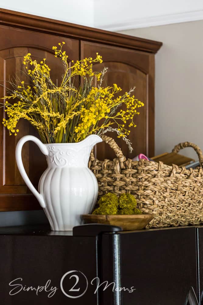Large white pitcher on top of a refrigerator filled with yellow fall flowers and dried grasses next to a large woven basket and a small wood bowl with moss-covered orbs.