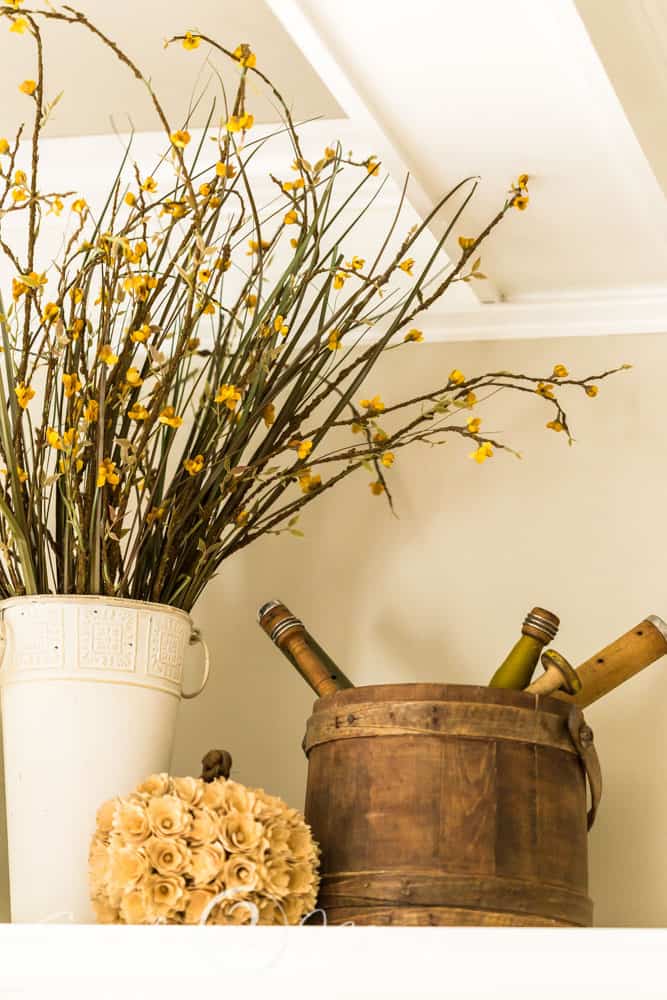 A tall painted tin bucket holds tall fall floral decor stems and sits next to a vintage wood firken filled with antique wooden spools. A white textured pumpkin sits in between.