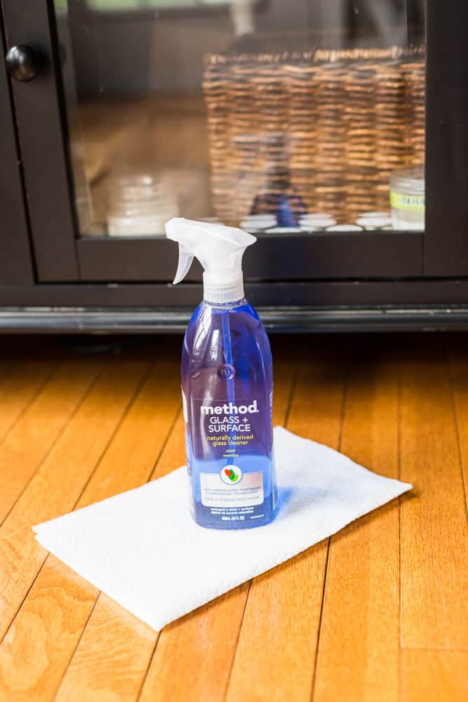 bottle of window cleaner sitting on paper towels on a wood floor in front of a cabinet with a glass door front