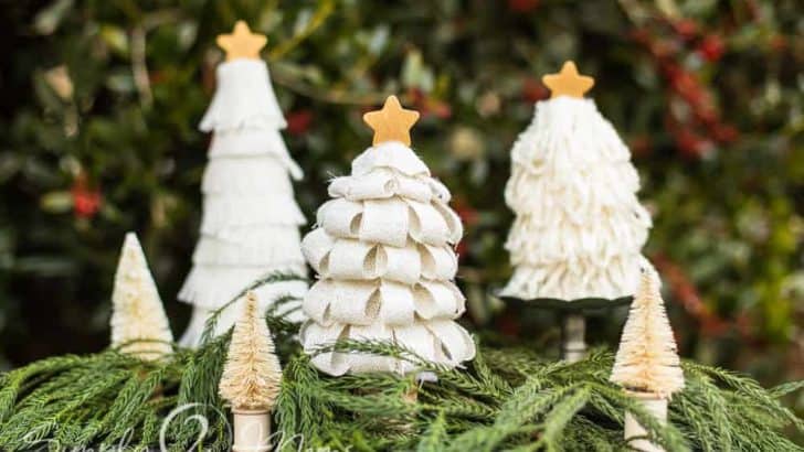 Burlap loop Christmas tree grouped with other neutral burlap, yarn, and bottle brush trees on a bed of fresh greenery with holly trees in the background.