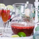 clear glass pitcher of Raspberry Prosecco cocktail on a white tray with champagne flutes filled with the cocktail and garnished with limes