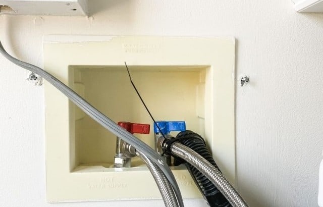 Uncovered washer hookup box in a laundry room with visible knobs for hot and cold water and three hoses coming out of it.