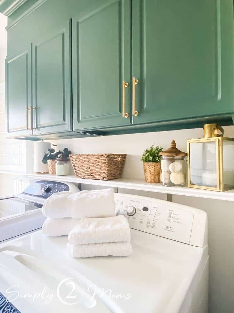 Laundry room with painted laminate cabinets over the washer and dryer and a shelf over washer and dryer with laundry room essentials and faux plants and baskets.