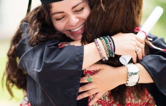 college graduate wearing cap and gown holding diploma in hand hugging woman