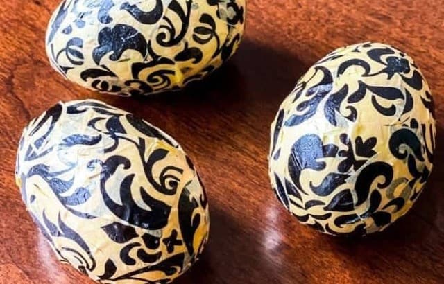 Three plastic eggs with black filigree decoupage pattern on a table with a potted plant in a basket container.