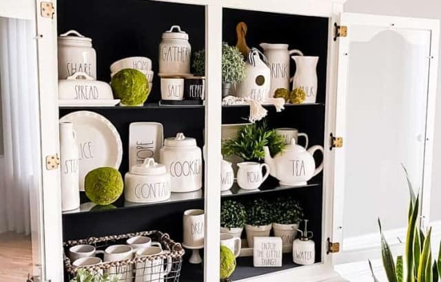 china cabinet with both hutch doors open and plant on stool in front of one door stoneware and greenery with faux plants and moss spheres styled on glass shelves