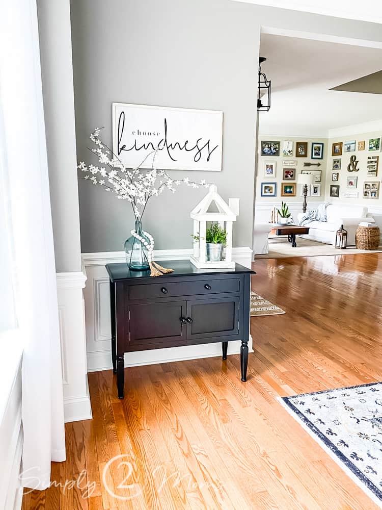 small side table with 2 drawers and double cabinet below. choose kindness word artwork hung on wall above table. glass jar with white floral stems and wood bead around jar and wood lantern with faux plant inside on top of table. living room beyond dining room can be seen in photo