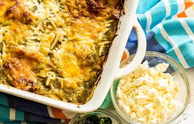 Rectangular casserole dish of Green Chile Chicken Enchiladas on a striped dishtowel next to small bowls filled with feta cheese, chopped cilantro, and diced red onion