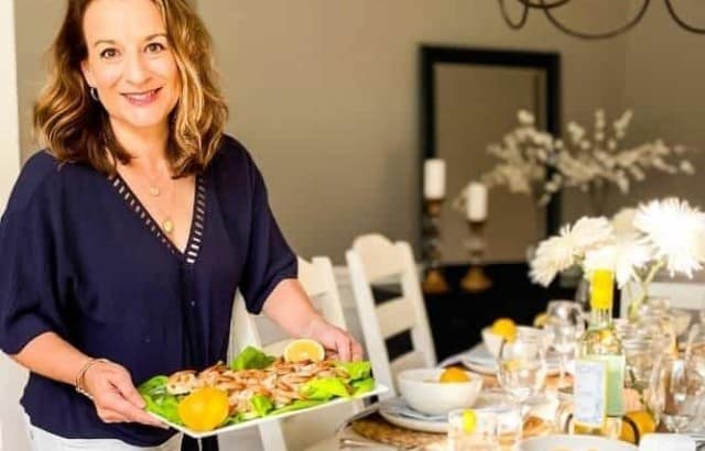Woman holding a platter of grilled shrimp next to a dining room table decorated for a summer dinner party with white dishes and lemons