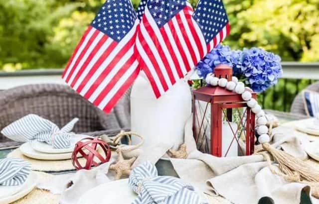 outdoor tablescape patriotic centerpiece decorated with vase filled with American flags, candle lantern, and vase of hydrangea on galvanized tray in middle of table outside