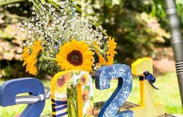 Paper mache numbers decorated with patterned scrapbook paper, paint and ribbon display the graduation year on a table in front of a vase of sunflowers