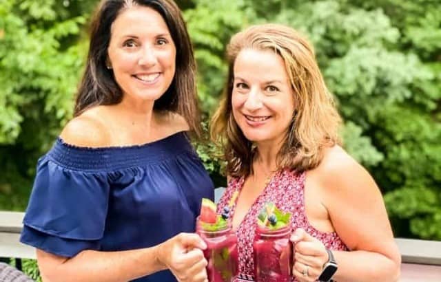 two woman holding glass mugs with fruit cocktail in them outside on deck