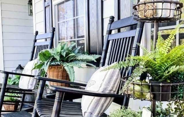 Front porch with black rocking chairs with neutral striped pillows and a large tiered plant stand filled with ferns and topiary plants.
