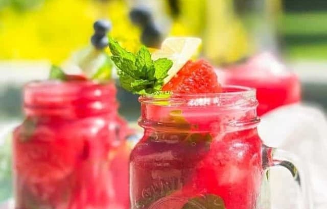 Two mason jar mugs filled with Watermelon Blueberry Mojitos on a tray with a carafe of more of the drink sitting outside by a pool