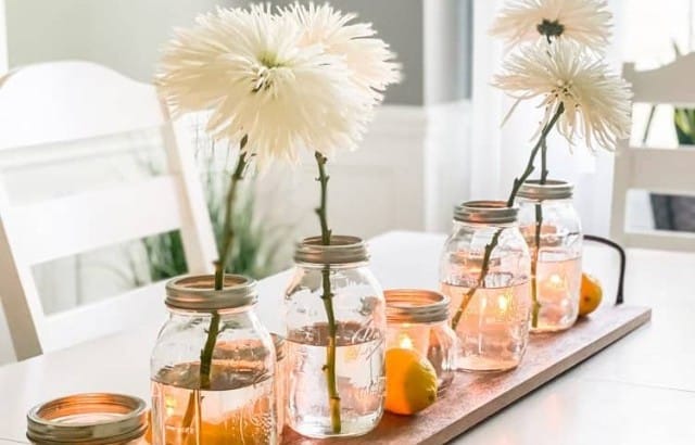 styled long slim wood tray with handles in middle of dining room table with 4 mason jar vases with one stem in each and other mason jars with candles and lemons in between flowers