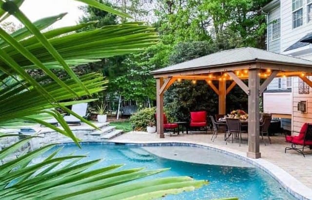 Costco cedar gazebo on a patio next to a pool with a house behind it.