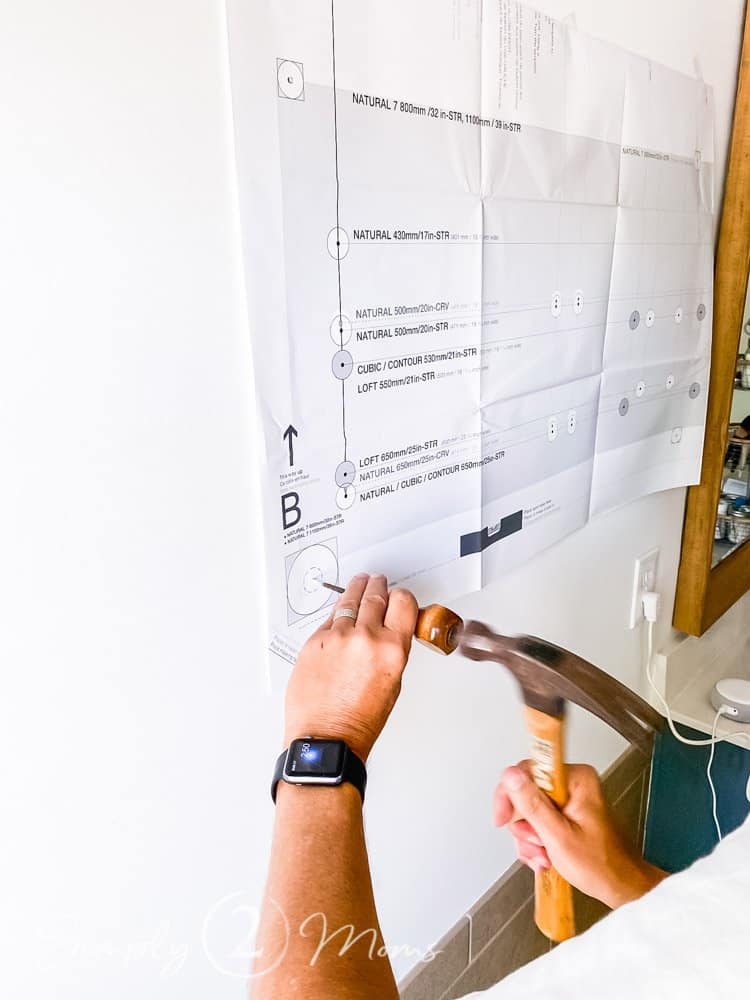 man's hands using awl and hammer to make hole in drywall behind paper template to install heated towel rack