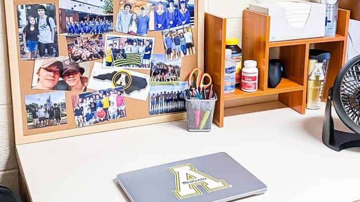 view of student desk in college dorm room with laptop and bulletin board leaning against wall filled with photos and a desk organizer on side of board with pencil cup on desk