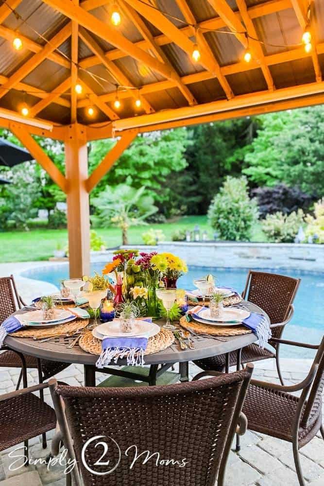 outdoor table set with flowers and place settings on patio under pergola with pool behind patio