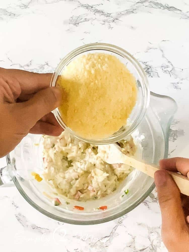 Hand holding a small bowl of grated Parmesan cheese over a clear glass mixing bowl of corn dip