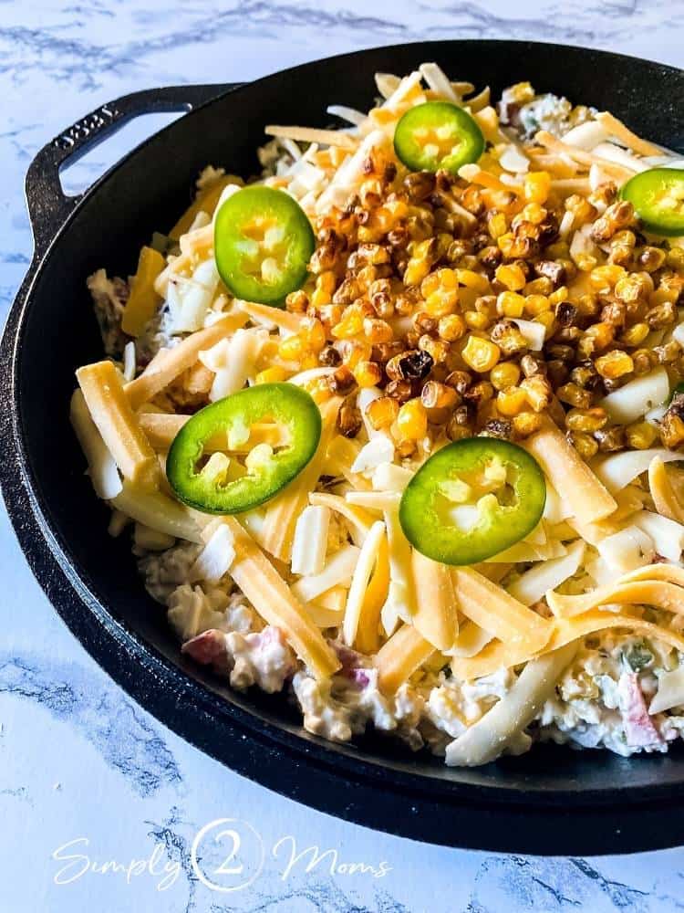 Cast iron skillet of Hot Roasted Corn Dip before being baked. Dip is topped with shredded cheese, sliced jalapeno peppers, and roasted corn.