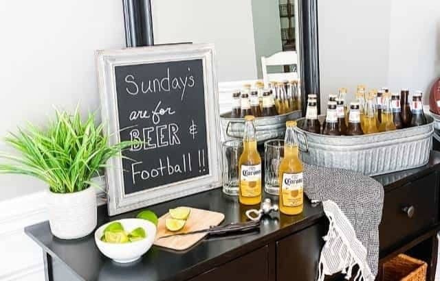 console table with mirror above and baskets in cubbies at bottom with galvanized tub of beer bottles, limes, faux plant and chalkboard with football saying written on it on top of table