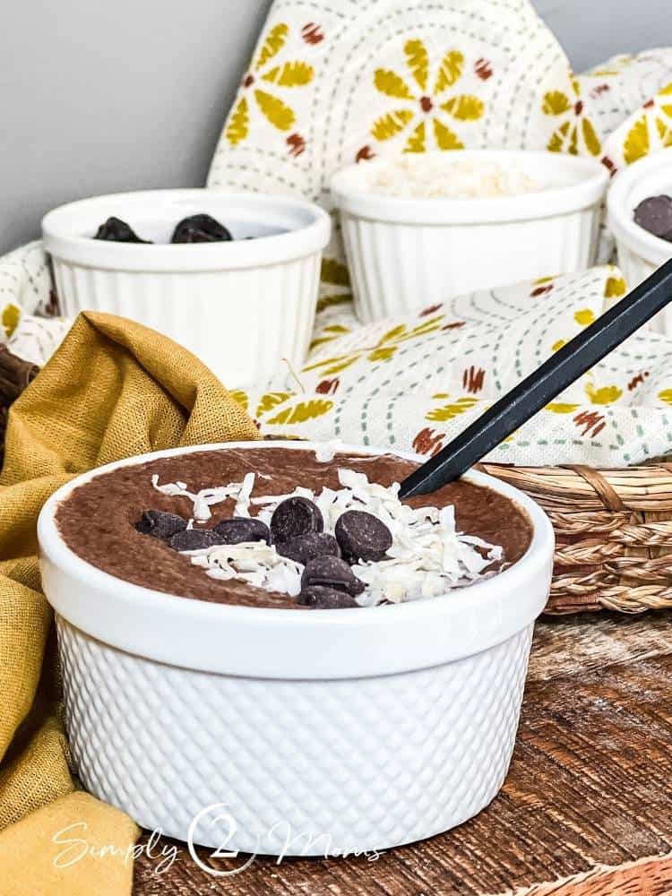 Cup of Mexican Chocolate Chia Pudding topped with coconut shreds and sugar free chocolate chips with cups of toppings in background