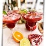 Two Pomegranate Margaritas on a table with cut pomegranate, orange, and lime