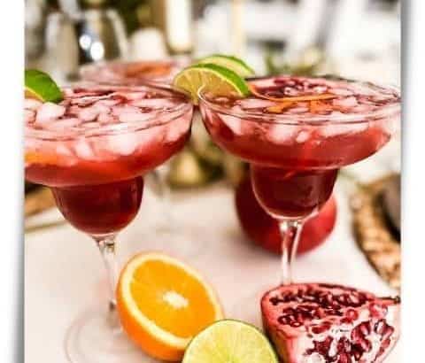 Two Pomegranate Margaritas on a table with cut pomegranate, orange, and lime