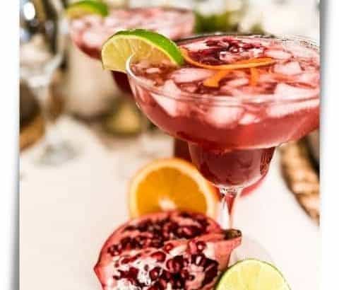 Pomegranate Margaritas on a table with cut pomegranate, orange, and lime