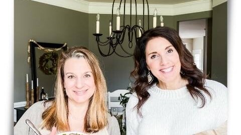 Two bloggers holding a wood bowl with fresh rolls and a serving dish of Brussel Sprouts for an Italian themed dinner party
