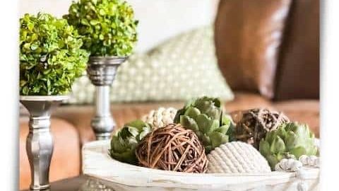 Coffee table with pewter candlesticks topped with boxwood urns next to a white dough bowl filled with artichokes and textured orbs.
