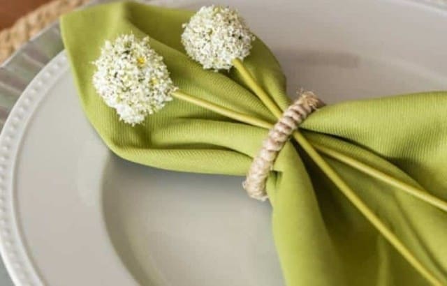 Jute placemat with silver charger & white plate with green cloth napkins held by jute napkin rings with white faux flowers.