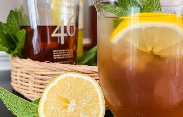 Large clear glass mug filled with Sweet Tea Bourbon cocktail on a tray with fresh mint and lemons & a bottle of whiskey