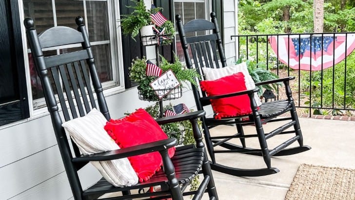 Two black rocking chairs on a front porch with patriotic throw pillows and bunting on the railing.