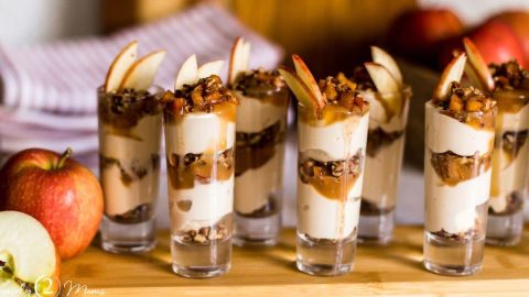 Shot glasses filled with keto caramel apple cheesecake parfaits on a wood board with whole apples.