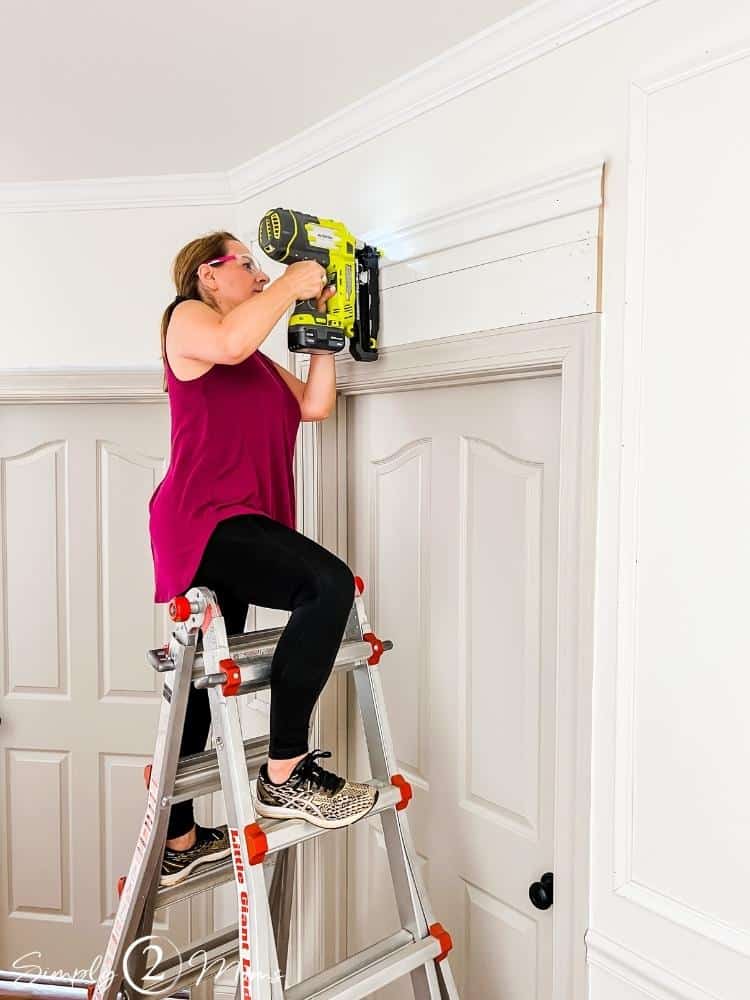 Woman on a ladder using a nail gun to attach trim to make a decorative door header.