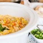 White crockpot insert filled with healthy crab and corn chowder on a buffet with a bowl of sliced green onions and basket of sliced baguettes.