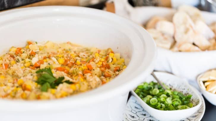White crockpot insert filled with healthy crab and corn chowder on a buffet with a bowl of sliced green onions and basket of sliced baguettes.