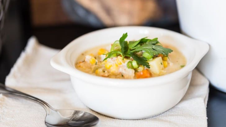Stoneware soup crock filled with healthy corn chowder with crab and a spoon beside the bowl.