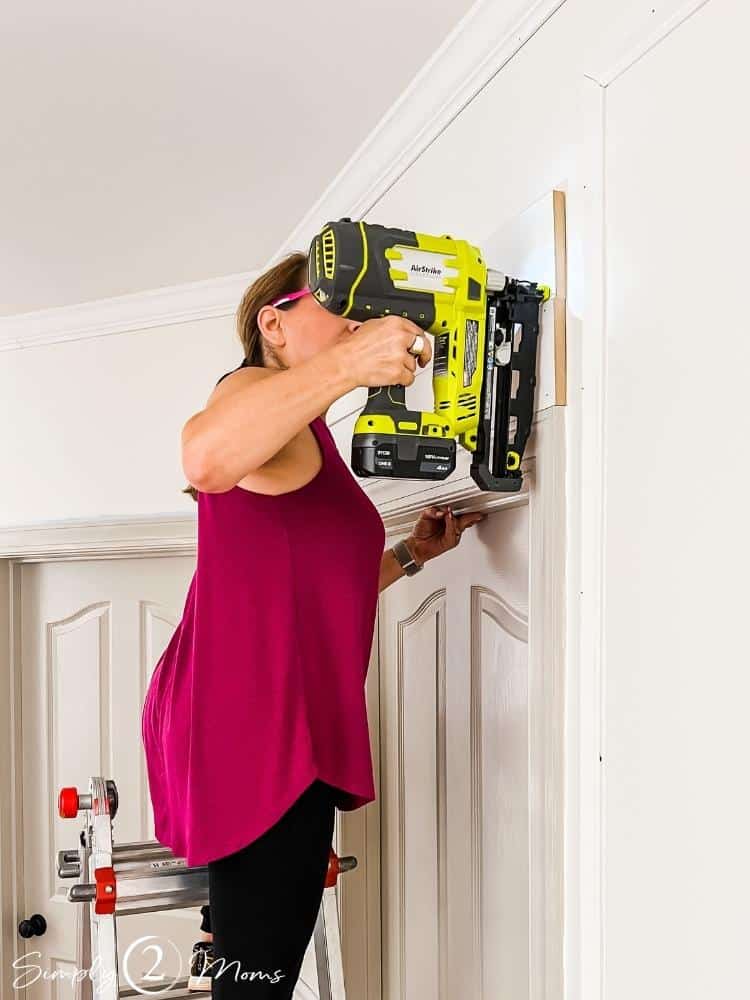 Woman on a ladder using a nail gun to attach stacked boards above an interior door frame to make a decorative header.
