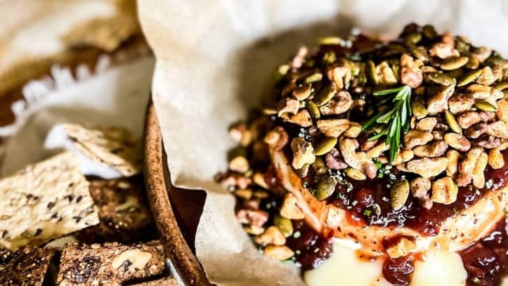 Round of Brie cheese topped with cranberry compote, toasted walnuts, pepitas, and rosemary on a parchment lined baking dish.