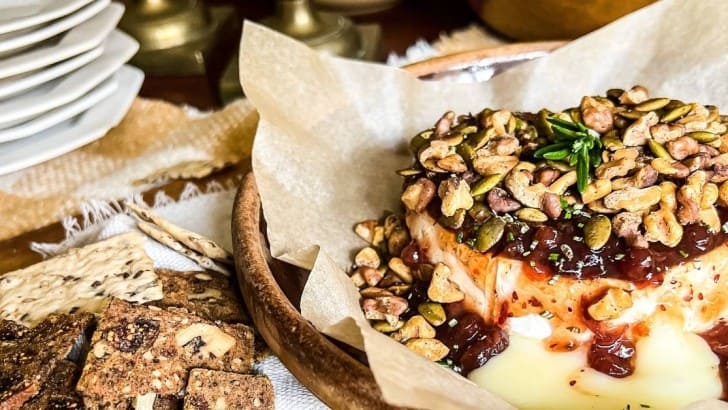 Round of Brie cheese topped with cranberry compote, toasted walnuts, pepitas, and rosemary on a parchment lined baking dish.